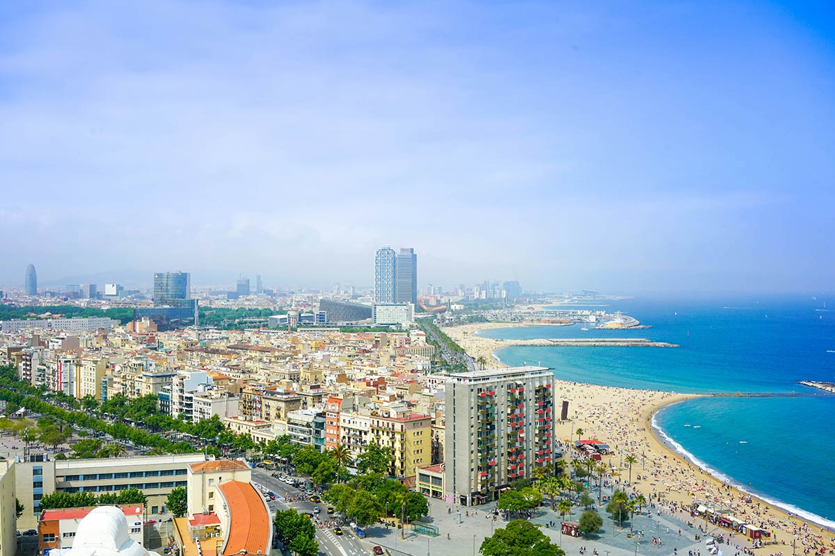 På bildet er det avbildet en panoramautsikt over Barcelona, Spania, med byens tettbygde områder som strekker seg mot den brede sandstranden og det glitrende Middelhavet. Dette er et populært reisemål for mange som vurderer å flytte til Spania.